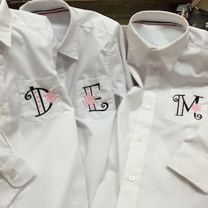 May include: Three white long-sleeve shirts with embroidered pockets. The pockets have a pink flower and a black letter, D, E, and M.