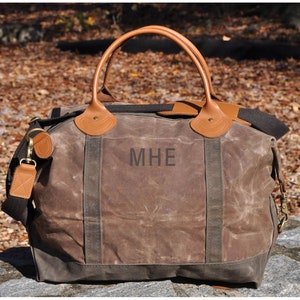 May include: A brown canvas and leather duffel bag with brown leather handles and a black shoulder strap. The bag has a monogram "MHE" on the front.