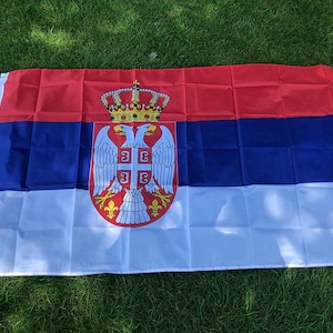 May include: A rectangular Serbian flag, featuring horizontal stripes of red, blue, and white. The Serbian coat of arms, including a crown, double-headed eagle, and shield, is centered on the blue stripe. The flag is displayed on green grass.