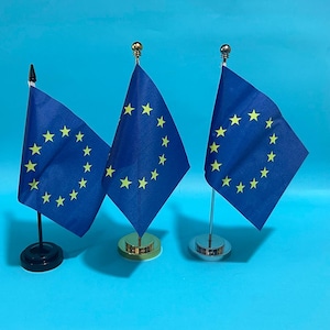 Puede incluir: Tres banderas en miniatura de la Unión Europea sobre soportes de escritorio individuales. Cada bandera es azul con un círculo de doce estrellas amarillas. Los soportes tienen acabados diferentes: negro, dorado y plateado, cada uno con un remate.