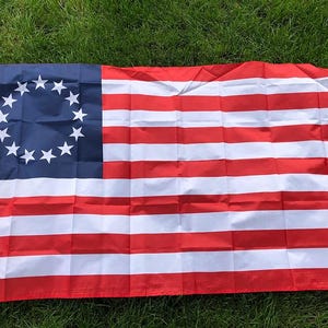 Könnte beinhalten: Eine Betsy-Ross-Flagge mit einem blauen Kanton mit 13 weißen Sternen in einem Kreis und 13 abwechselnd roten und weißen Streifen. Die Flagge besteht aus leichtem Material und wird im Freien auf einer grasbewachsenen Oberfläche gezeigt.
