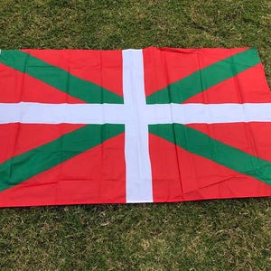 May include: A rectangular flag with a red background, featuring a white cross and green diagonal stripes. The flag is lying on a bed of green grass. The flag has a white border with grommets.