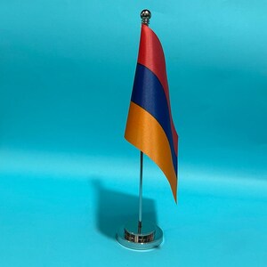 Puede incluir: Una pequeña bandera de escritorio de Armenia con franjas horizontales de color rojo, azul y naranja. La bandera está unida a un poste de metal vertical delgado de color plateado montado sobre una base circular de metal plateado pulido.