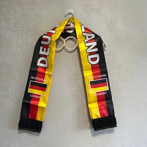 Könnte beinhalten: Ein Schal mit Satin-Finish in den Farben Schwarz, Rot und Gold der deutschen Flagge. Das Wort DEUTSCHLAND ist in großen weißen Buchstaben gedruckt, mit dem Text DEUTSCHLAND und GERMANY unter kleinen Flaggen an beiden Enden mit schwarzer Franse.