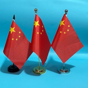 May include: Three miniature flags of China, each featuring a red field with five yellow stars. The flags are mounted on individual stands: one with a black base and finial, one with a gold-tone base and finial, and one with a silver-tone base and finial.