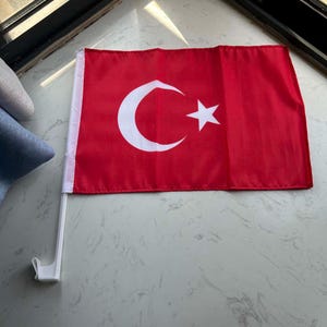 Op de afbeelding: Een rechthoekige rode vlag met een witte halve maan en een vijfpuntige ster in het midden. De vlag is bevestigd aan een witte plastic stok met een haak aan de onderkant, ontworpen voor montage op een voertuigruit of vergelijkbaar oppervlak.