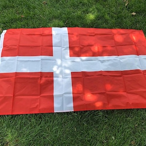 Könnte beinhalten: Eine rote Flagge mit einem weißen skandinavischen Kreuz-Design, das auf grünem Gras liegt. Das rote Feld der Flagge wird von einem weißen Kreuz geteilt, ein Symbol der nationalen Identität.