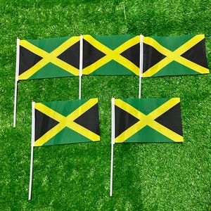 Op de afbeelding: Een verzameling van vijf kleine handvlaggen van Jamaica, elk voorzien van een geel diagonaal kruis dat de stof in vier zwarte en groene driehoeken verdeelt. Elke vlag is bevestigd aan een dunne witte plastic stok met een kleine gouden dop.