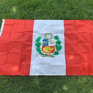 May include: A rectangular Peruvian flag with a central white stripe flanked by red stripes. The flag features the Peruvian coat of arms in the center, including a vicuña, a cinchona tree, and a cornucopia. The flag is lying on green grass.