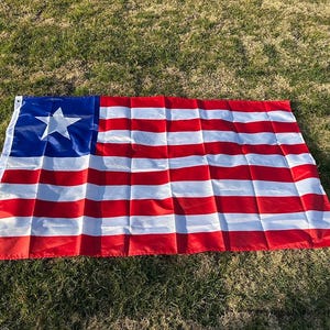 May include: A rectangular flag with a blue canton featuring a white star. The flag has alternating red and white horizontal stripes. The flag is lying on a bed of green grass.