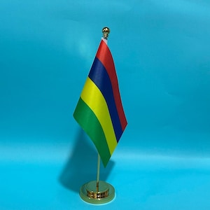 Puede incluir: Una pequeña bandera de escritorio de Mauricio con franjas horizontales de color rojo, azul, amarillo y verde. La bandera está sujeta a un fino mástil de metal dorado con una base redonda y un remate esférico dorado en la parte superior.