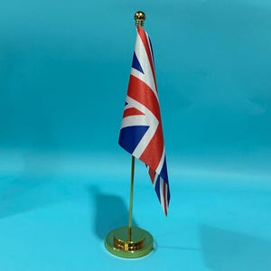 Puede incluir: Una pequeña bandera del Reino Unido unida a un poste de metal vertical y delgado de color dorado. La bandera presenta el diseño tradicional de cruz roja, blanca y azul. El poste está montado sobre una base circular dorada con un remate esférico.
