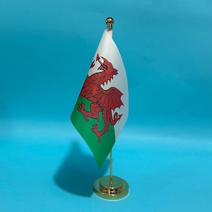 Puede incluir: Una pequeña bandera de escritorio de Gales con un fondo horizontal blanco y verde y un gráfico de un gran dragón rojo. La bandera está unida a un fino mástil de metal dorado con una base redonda y un remate esférico dorado en la parte superior.