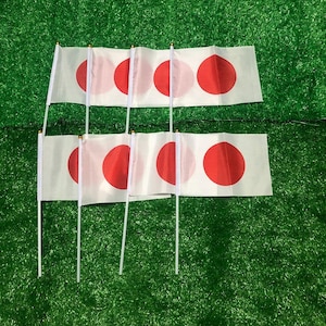 以下が含まれることがあります： 白い長方形の地に中央に赤い円が描かれた、6本の手持ち旗のセット。各旗は、先端に小さな金色のビーズが付いた細い白いプラスチック製の棒に取り付けられており、日本の国旗を表現しています。