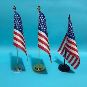 May include: Three miniature United States flags on stands. The first has a silver pole and base, the second a gold pole and base, and the third a black pole and base. Each flag features the traditional red, white, and blue star-spangled design.