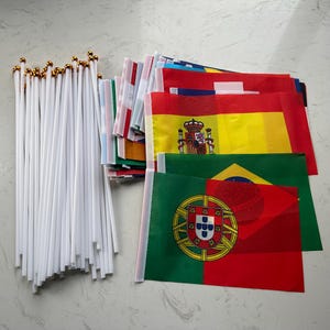 May include: A collection of small rectangular national flags attached to thin white plastic poles with gold-colored spherical finials. The flags feature various colorful designs, including the national emblems of Portugal, Brazil, and Spain, stacked together.