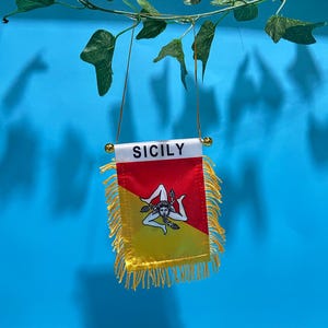 Peut inclure: Un petit fanion rectangulaire portant le mot SICILY en lettres noires sur une bande blanche. Le drapeau est divisé en diagonale en sections rouge et jaune, avec un emblème central triskèle blanc et une bordure à franges dorées.