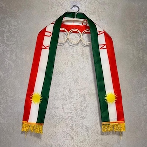May include: A long, rectangular sash featuring vertical stripes of red, white, and green. The text "KURDISTAN" is printed in red on the white stripe. Each end displays a yellow sun emblem with rays and is finished with yellow fringe trim.