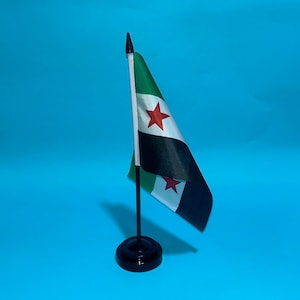 Puede incluir: Una pequeña bandera de escritorio con franjas horizontales verde, blanca y negra, con dos estrellas rojas centradas en la franja blanca. La bandera está sujeta a un mástil blanco fino con un remate negro, sobre una base redonda negra.