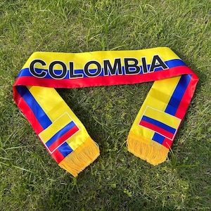Può includere: Una sciarpa in raso con le strisce orizzontali gialla, blu e rossa della bandiera colombiana. La parola COLOMBIA è stampata in grandi lettere maiuscole nere sulla parte superiore, con icone di bandiere più piccole e frange gialle.