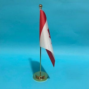 Op de afbeelding: Een kleine Canadese vlag bevestigd aan een dunne, verticale goudkleurige metalen paal met een ronde, bijpassende goudkleurige voet. De vlag heeft het iconische rood-witte ontwerp met een centraal rood esdoornblad op een lichtblauwe achtergrond.