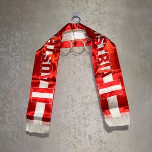 Könnte beinhalten: Ein rot-weißer Satinschal mit dem Schriftzug AUSTRIA in fetten weißen Großbuchstaben auf beiden Seiten. Der Schal zeigt das Design der österreichischen Flagge und ist an beiden Enden mit weißen Fransen versehen, hängend an einem Kleiderbügel.