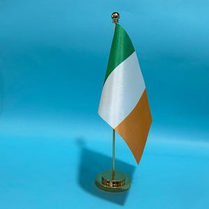 May include: A small desk flag of Ireland featuring vertical stripes of green, white, and orange. The flag is attached to a thin, vertical gold-colored metal pole that is mounted on a circular, polished gold-colored metal base with a small ball finial.