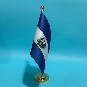 Op de afbeelding: Een kleine bureuvlag van El Salvador met twee horizontale blauwe banen gescheiden door een witte baan in het midden. Het nationale wapenschild staat centraal op de witte baan. De vlag is bevestigd aan een goudkleurige metalen stok en voet.