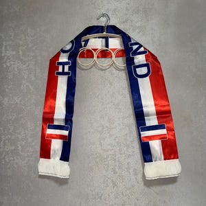 May include: A satin scarf featuring vertical stripes in blue, white, and red. The text HOLLAND is printed in blue capital letters along the fabric. Each end of the scarf is finished with white fringe, and a small rectangular flag icon is visible.