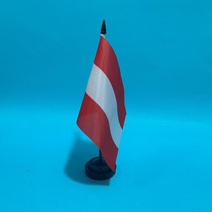 Puede incluir: Una pequeña bandera de escritorio con el diseño tricolor horizontal de Austria, que consta de dos franjas rojas separadas por una franja blanca central. La bandera está unida a un mástil negro fino con un remate puntiagudo, sobre una base redonda negra.