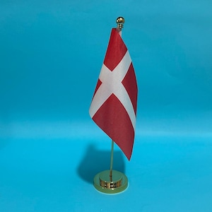Puede incluir: Una pequeña bandera de escritorio de Dinamarca que presenta un campo rojo con una cruz nórdica blanca. La bandera está unida a un poste de metal vertical delgado de color dorado montado sobre una base circular de metal dorado pulido con un remate dorado.