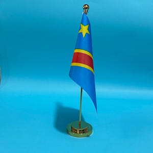 May include: A small desk flag of the Democratic Republic of the Congo featuring a light blue field with a yellow star in the upper left corner and a diagonal red stripe bordered by yellow. The flag is mounted on a gold-colored metal pole and base.