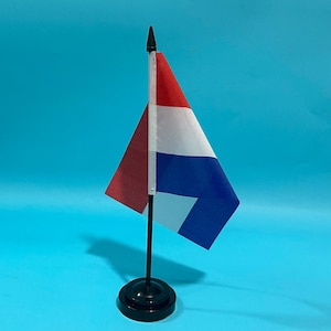 Op de afbeelding: Een kleine bureuvlag met horizontale banen in rood, wit en blauw, bevestigd aan een dunne zwarte plastic stok met een puntige top. De vlag is gemonteerd op een ronde zwarte voet en toont de nationale kleuren van Nederland.