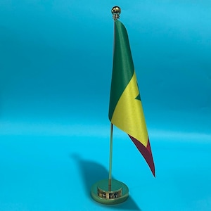 Puede incluir: Una pequeña bandera de escritorio de Senegal con franjas verticales verde, amarilla y roja y una estrella verde central, sujeta a un fino mástil de metal dorado con base redonda y un remate esférico en la parte superior.