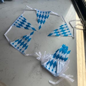 May include: A string of triangular pennant flags featuring a blue and white diamond pattern. The bunting is attached to a white fabric ribbon, with some sections of the flags spread out and others folded into a stack with visible white ribbon ties.