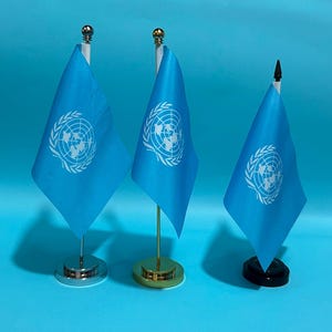 Op de afbeelding: Drie bureaustandaarden van de Verenigde Naties met een lichtblauw veld en een wit embleem. De vlaggen staan op verticale stokken met verschillende voetstukken: een zilveren, een gouden en een zwarte. Elke stok heeft een kleine decoratieve knop.