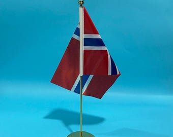 Może przedstawiać: Mała flaga biurkowa Norwegii z czerwonym tłem i niebieskim krzyżem skandynawskim z białymi obwódkami. Flaga jest przymocowana do pionowego białego masztu ze złotym zwieńczeniem na szczycie, osadzonego na okrągłej złotej metalowej podstawie.