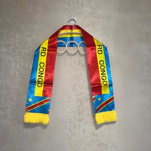 May include: A satin sash featuring the flag of the Democratic Republic of the Congo with vertical stripes of blue, yellow, and red. The text RD CONGO is printed in black on the yellow stripe, and the ends are finished with yellow fringe.