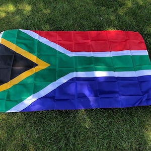 Könnte beinhalten: Eine südafrikanische Flagge mit einem lebendigen Design in den Farben Rot, Weiß, Grün, Blau, Schwarz und Gelb. Die rechteckige Flagge liegt auf grünem Gras und zeigt ihr unverwechselbares Muster.