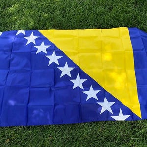 Könnte beinhalten: Rechteckige Flagge von Bosnien und Herzegowina mit blauem Feld, gelbem Dreieck und weißen Sternen. Die Flagge liegt auf grünem Gras. Die Abmessungen der Flagge sind sichtbar.