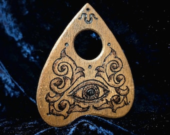 Puede incluir: Una planchette de tablero Ouija de madera con un diseño de ojo tallado y remolinos intrincados. La planchette tiene forma de lágrima y tiene un agujero en el centro.