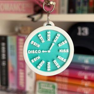 May include: A round keychain with a teal face and white border. The face has the word "DISCO" and the word "KISS" repeated around the edge. The keychain is hanging in front of a bookshelf filled with books.