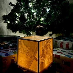 May include: A lit-up, square, wooden lamp with engraved images of people, sitting in front of a Christmas tree with ornaments and lights. Wrapped gifts are in the foreground. The lamp emits a warm, inviting glow.