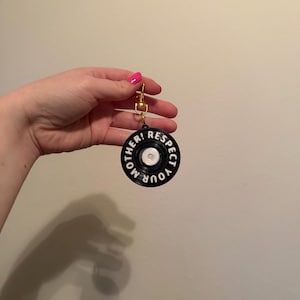 May include: Black and white circular keychain with the text "RESPECT YOUR MOTHER!". The keychain has a gold-colored clasp and a white center. The text is in a bold, sans-serif font.