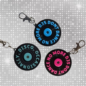 May include: Three black vinyl record keychains with silver clasps. Each keychain has a different colored center and text: blue, "MORE DJS DON'T DANCE ON"; teal, "DISCO, OCCASIONALLY"; and pink, "DANCE NO MORE DJS DON'T".