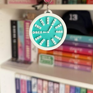 May include: A round keychain with a teal face and white border. The face has the word "DISCO" and the word "KISS" repeated around the edge. The keychain is hanging in front of a bookshelf filled with books.