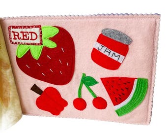 Puede incluir: Una página de libro de fieltro rosa con frutas rojas de fieltro y un tarro de mermelada. La palabra "RED" se muestra encima de una fresa roja con hojas verdes. Otras frutas de fieltro incluyen cerezas, una manzana y una rodaja de sandía.