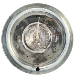 May include: A silver-colored clock with a circular face. The clock features a central emblem and radial lines. The numbers 12, 3, 6, and 9 are visible. The clock hands are white.