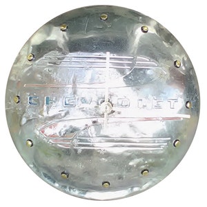May include: A round, silver-colored metal clock with the word "CHEVROLET" embossed on the face. The clock has a vintage design with a classic car emblem and small gold-colored rivets around the edge.