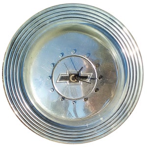 May include: A round, silver-colored clock made from a car wheel cover. The clock face features the Chevrolet logo in the center, with black clock hands. The wheel cover has a series of concentric ridges around the edge.